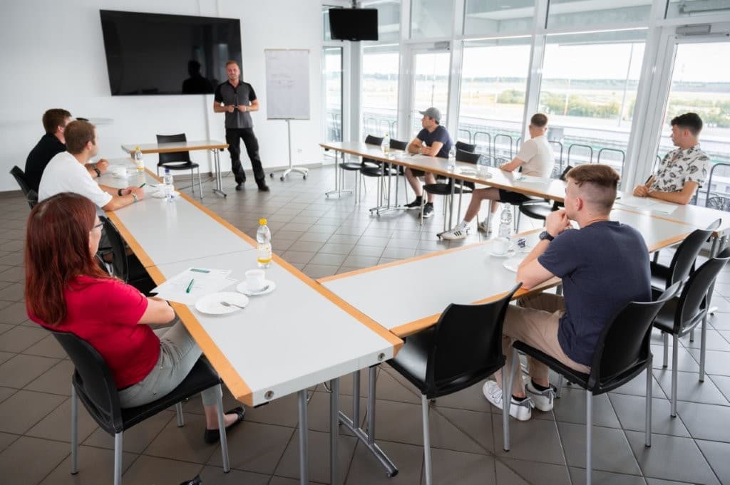 Theorieteil im Schulungsraum: Trainer am Flipchart, Teilnehmende an U-förmig gestellten Tischen, Panoramafenster mit Blick auf die Übungsstrecke